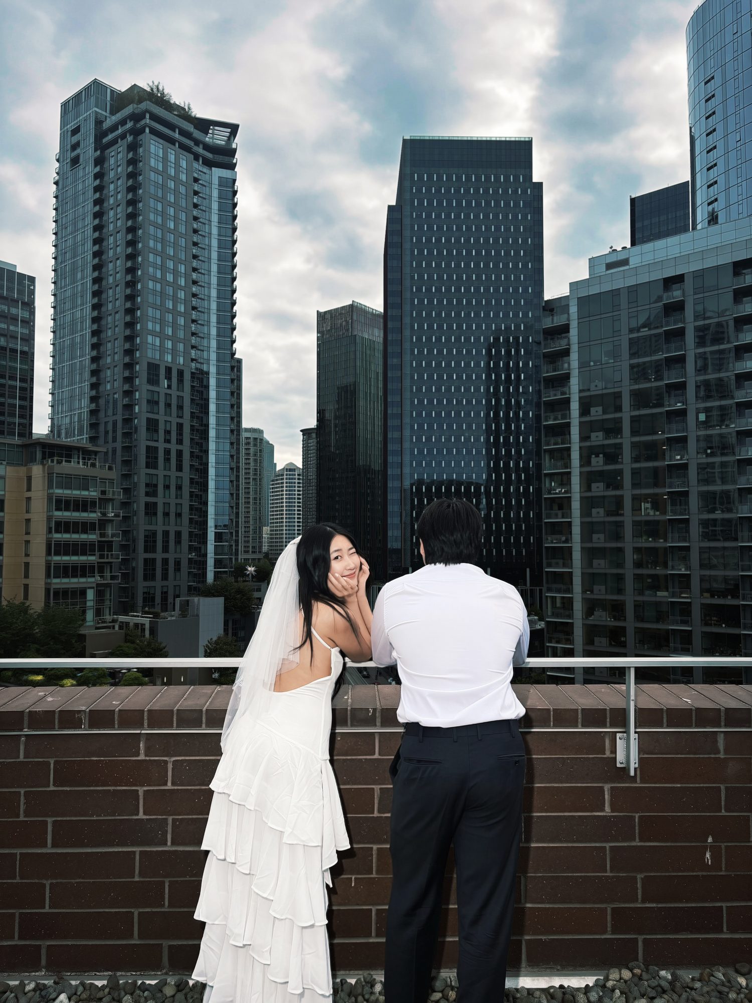 Lucia, in a white tiered dress and veil, turns back from a rooftop railing and winks at the camera. Teddy stands beside her facing the Seattle skyline.